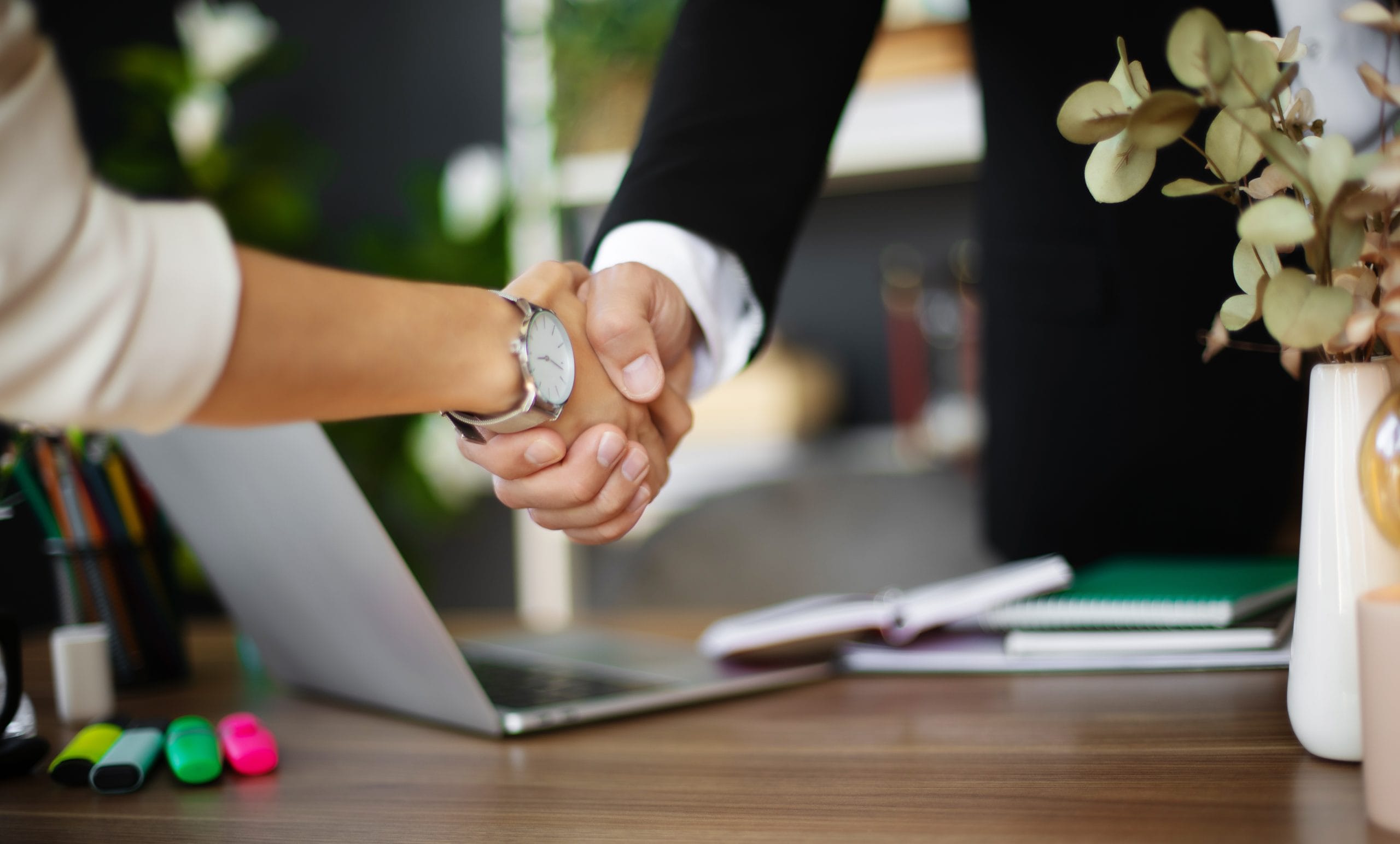Two individuals shake hands over a desk with a laptop, office supplies, and a vase of flowers in the background. The image conveys a sense of professionalism, teamwork, and successful collaboration.
